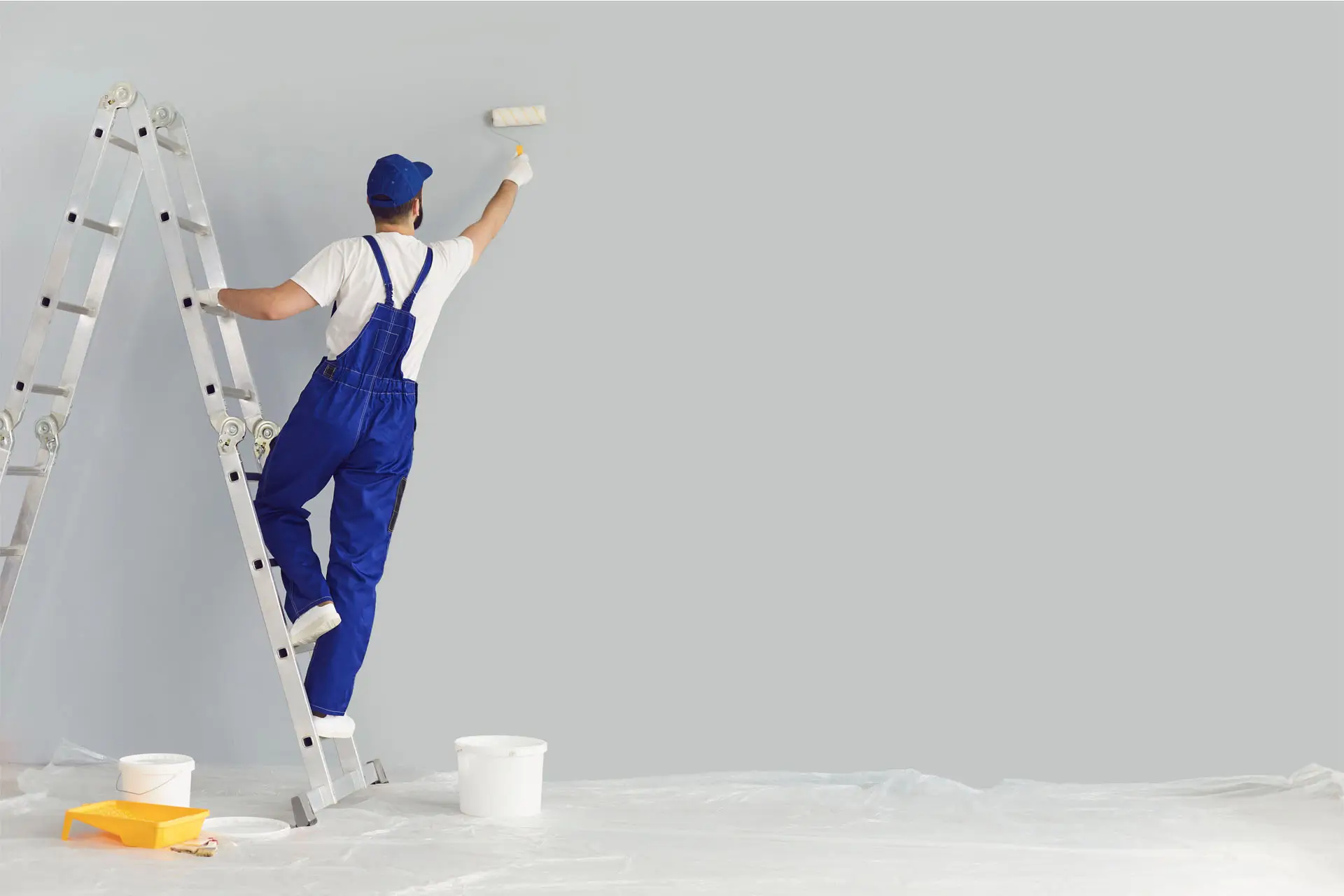 A man wearing overalls is painting a wall with a roller brush, focused on his task.